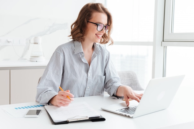 Happy brunette businesswoman in glasses using laptop computer while working in light apartment