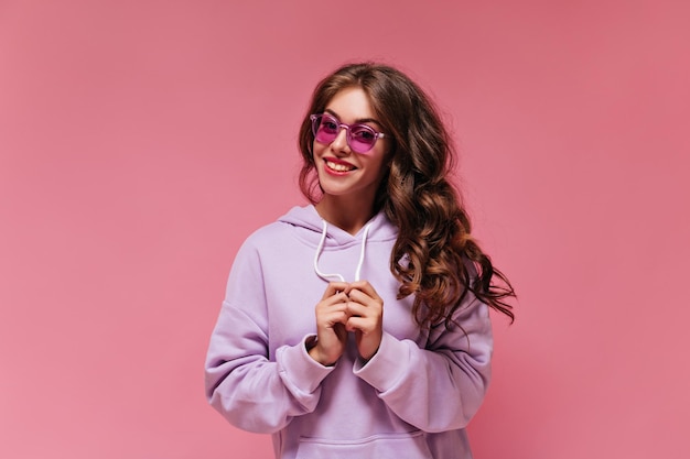 Happy brunette curly woman in purple oversized hoodie looks into camera Girl in bright sunglasses smiles on isolated pink background
