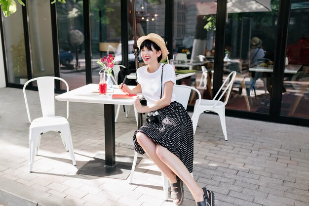 黒いスカートと白いシャツを着た幸せなブルネットの少女が屋外カフェに座って、良い気分で街の景色を楽しんでいます