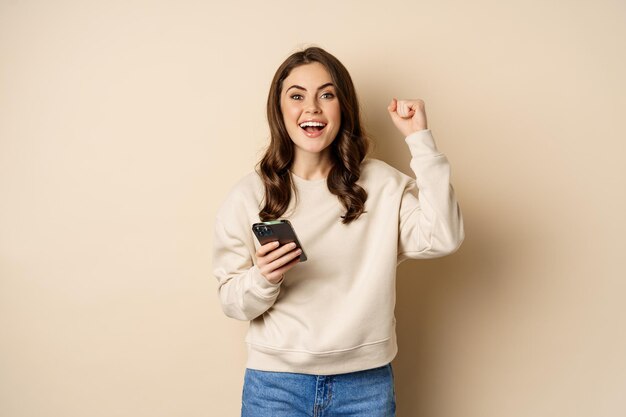 Happy brunette girl holding smartphone and cheering, winning, celebrating win on mobile phone app, standing over beige background.