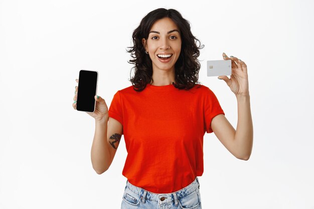 Счастливая брюнетка улыбается, рекомендует приложение или банк, показывает приложение для покупок на пустом экране смартфона, кредитную карту в руке, белый фон.