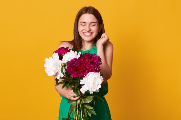 happy brunette girl, stands with smiling and holding bouquet of white and burgundy peonies, expresses happyness and gladness, has gift for holiday, looks excited. People emotions concept.