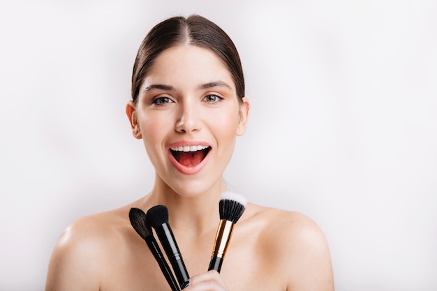 Happy brunette girl without makeup smiling, holding makeup brushes on white wall.