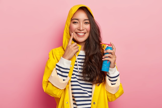 Happy brunette lady cures sore throat with spray, dressed in yellow raincoat with hoody, being ill after spending long time outdoor during rainy day