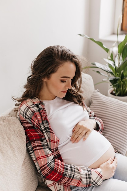 Happy brunette pregnant woman in stylish plaid shirt smiles and gently touches belly Cheerful mother in white tee chilling out on beige sofa