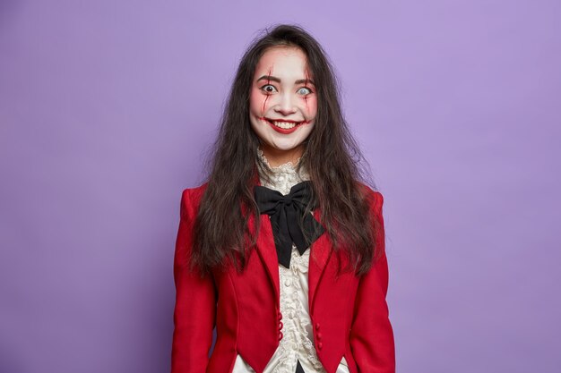 Happy brunette terrifying Asian woman has spooky eyes with lenses and blood scars dressed in masquerade costume smiles positively isolated on vivid purple wall