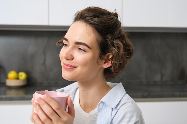 Foto gratuita una donna bruna felice che beve caffè a casa una ragazza con una tazza rosa si siede nella sua cucina e si diverte