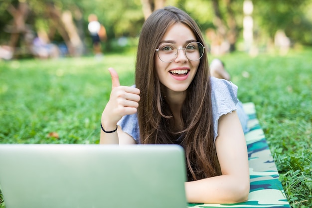 Счастливая брюнетка женщина в очках, лежа на траве в парке с портативным компьютером и показывая большой палец вверх