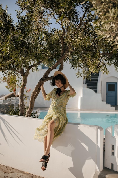 Happy brunette woman in hat and green dress sits in white fence and smiles Pretty shorthaired lady in boater poses in good mood near olive tree and pool