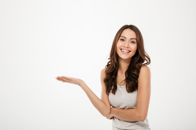 Happy brunette woman holding copyspace on the palm and looking at the camera over gray