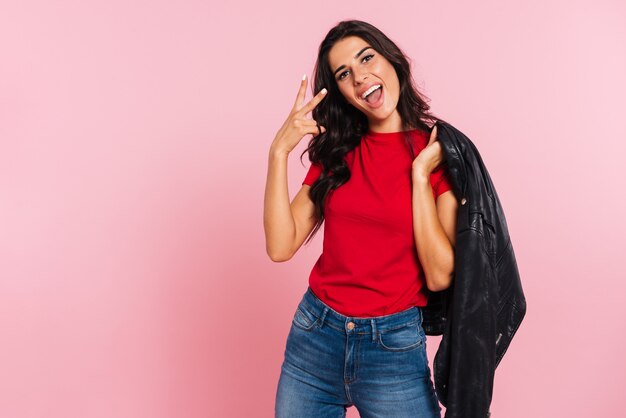 Happy brunette woman showing peace sign and looking at the camera while holding jacket on her shoulder over pink 