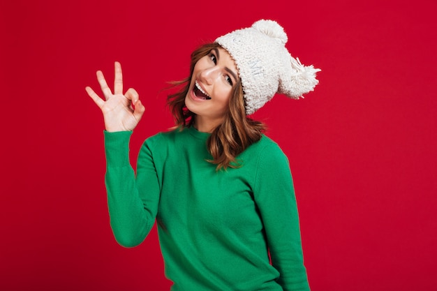 Happy brunette woman in sweater and funny hat showing ok