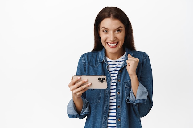 Happy brunette woman winning money on phone, playing mobile video game and rejoicing, celebrating goal achievement in app, standing against white background