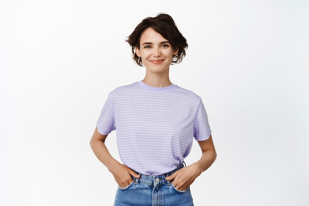 Happy brunette woman with short hair, standing in confident relaxed pose, hands in jeans pockets, smiling pleased, studio.