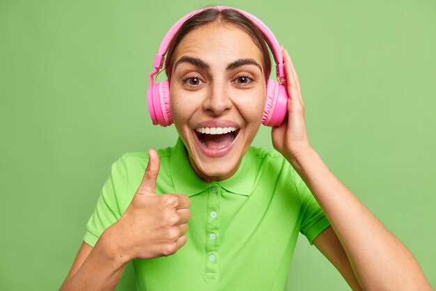 행복한 브루네트의 젊은 유럽 여성은 그녀의 재생 목록이 선명한 녹색 배경 위에 격리된 캐주얼한 티셔츠를 입은 귀에 분홍색 스테레오 헤드폰을 착용하도록 하는 재생 목록을 권장합니다.