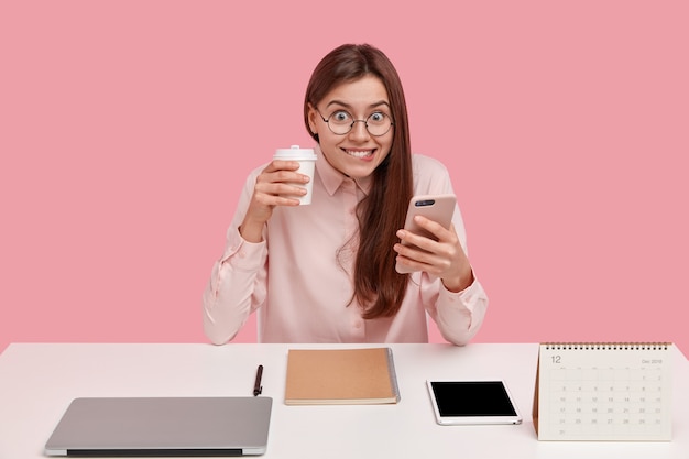 Happy brunette young woman holds modern cell phone, types text messages, carries takeaway coffee in hand, uses notepad for recording information