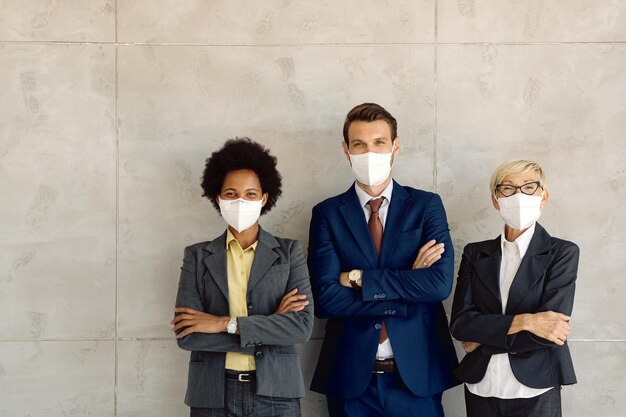 Happy business colleagues with face masks standing with crossed arms against the wall