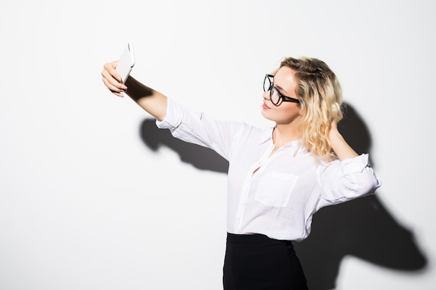 Happy business woman in eyeglasses taking selfie photo smartphone isolated on white wall