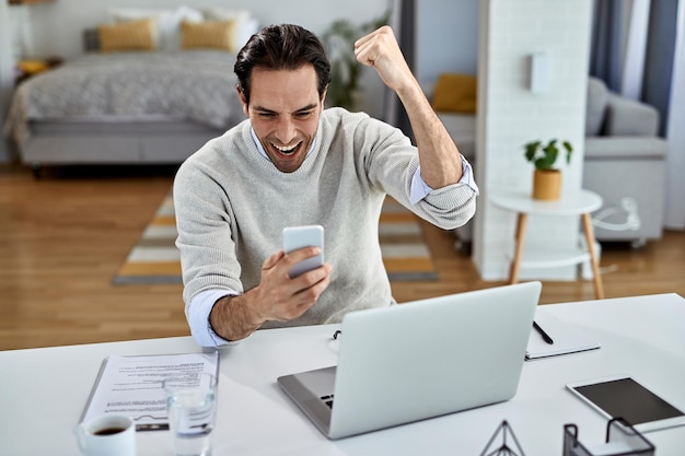 Happy businessman celebrating while reading good news on mobile phone at home