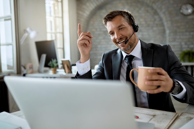 Foto gratuita uomo d'affari felice che beve caffè mentre effettua una videochiamata sul laptop in ufficio