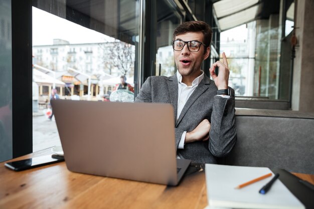 Happy businessman in eyeglasses sitting by the table in cafe with laptop computer and having idea