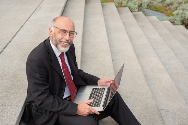 Happy businessman in eyeglasses using laptop on street