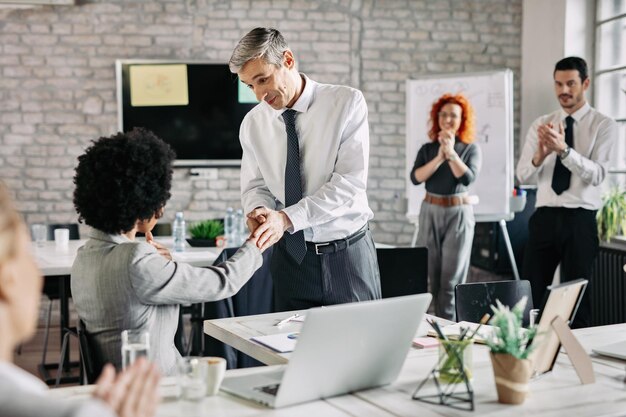 Happy businessman and his African American female colleague shaking hands after successful agreement in the office while other coworkers are applauding to them