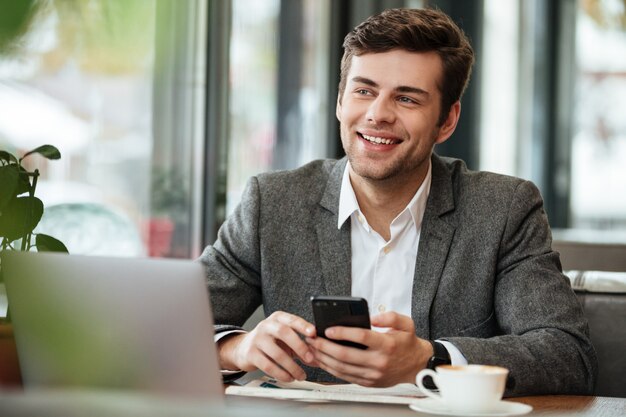 Happy businessman sitting by the table in cafe with laptop computer and smartphone while looking away