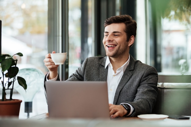 Happy businessman sitting by the table in cafe with laptop computer while holding cup of coffee and looking away