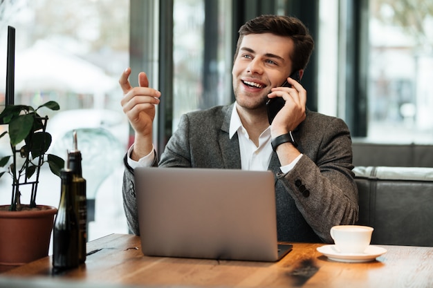 Happy businessman sitting by the table in cafe with laptop computer while talking by smartphone