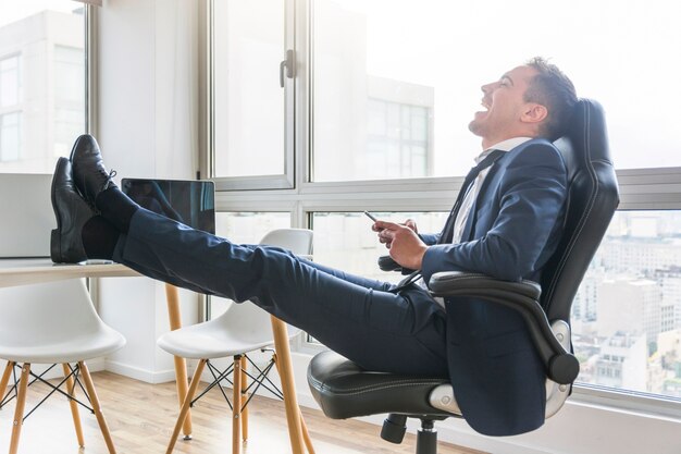 Happy businessman sitting on chair at workplace using mobile