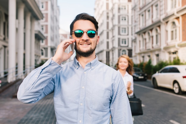 Happy businessman in sunglasses speaking on phone on street. Pretty blonde girl catching him from behind