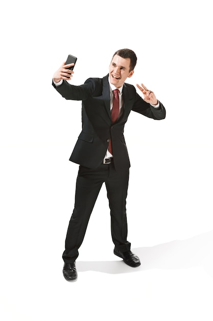 Счастливый бизнесмен разговаривает по телефону на белом фоне в студии съемки. Улыбающийся молодой человек в костюме стоит и делает селфи фото.