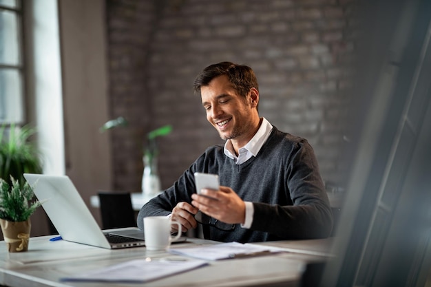 Free photo happy businessman texting on mobile phone while working on laptop in the office