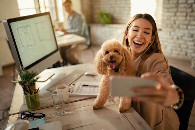 Happy businesswoman having fun while taking selfie with her dog in the office