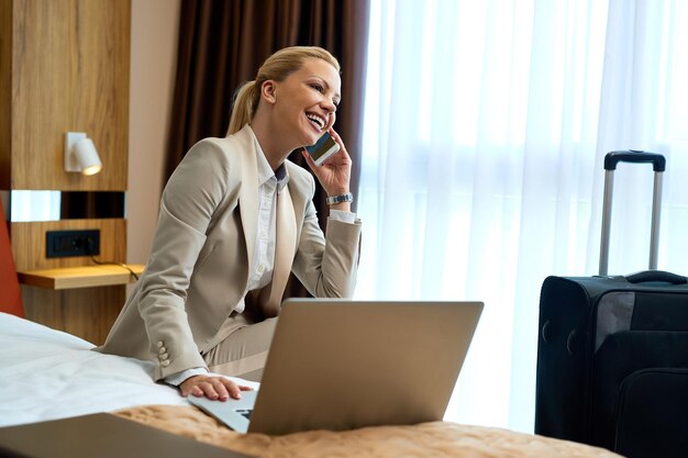 Happy businesswoman sitting on the bed in hotel room and talking on mobile phone