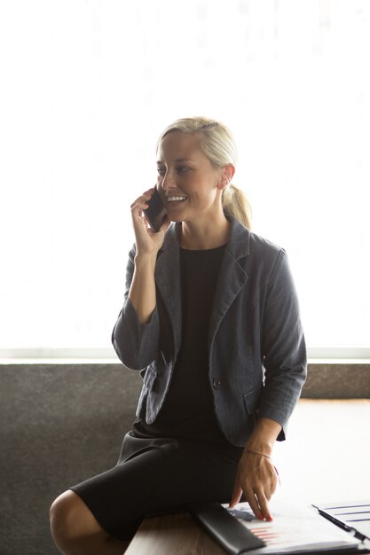 Happy businesswoman sitting on table and calling