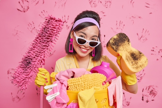 Happy busy Asian lady from cleaning service busy tidying up apartment equipped with sponge and mop stands dirty surrounded by pile of laundry wears headband sunglasses rubber gloves does home chores