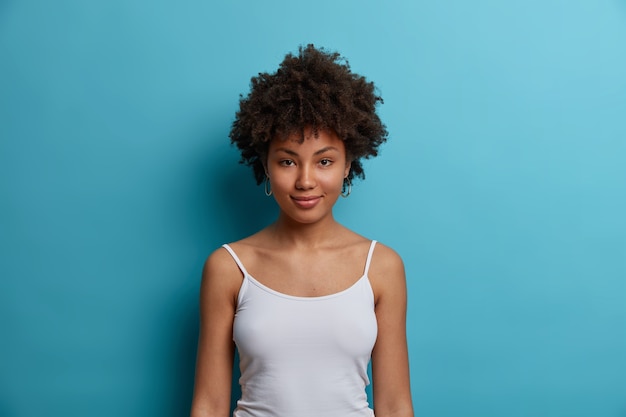 Happy calm dark skinned millennial girl spends lazy weekend at home, dressed in casual vest, feels confident and healthy, isolated on blue wall. People, lifestyle and emotions concept.