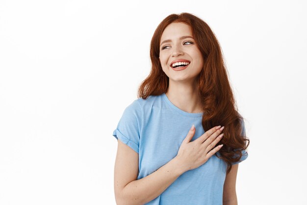 Happy candid redhead girl laughing and smiling naturally, holding hand on heart and looking aside at something beautiful and positive, reading logo, standing on white