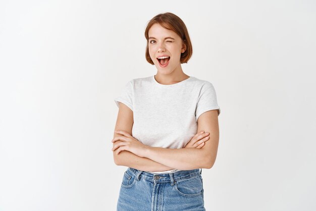 Happy candid woman in tshirt and jeans winking and smiling at camera motivating you cross arms on chest and look joyful standing on white background