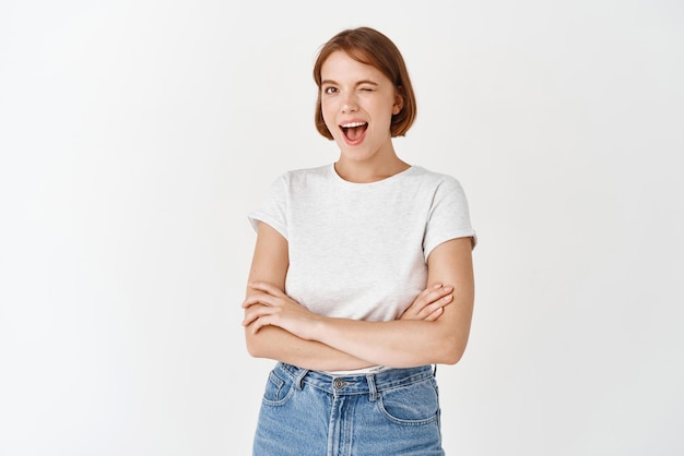 Happy candid woman in tshirt and jeans winking and smiling at camera motivating you cross arms on chest and look joyful standing on white background