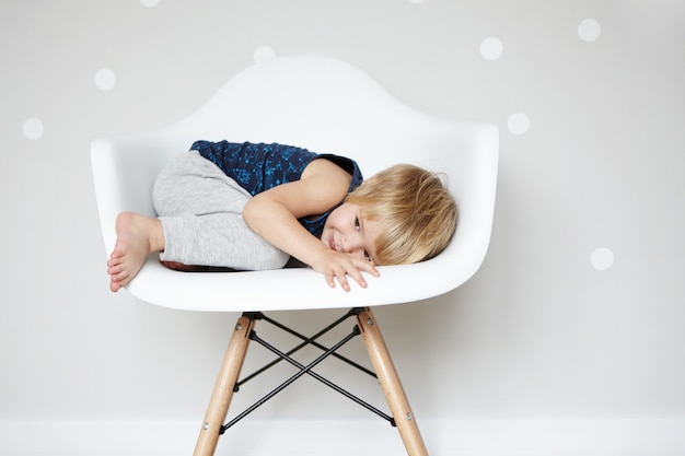 幸せな屈託のない子供時代。白人の幼児が白いデザイナーの椅子に身を包み込み、かくれんぼをしている間、友達から隠れています。室内で楽しんでいるかわいい男の子。