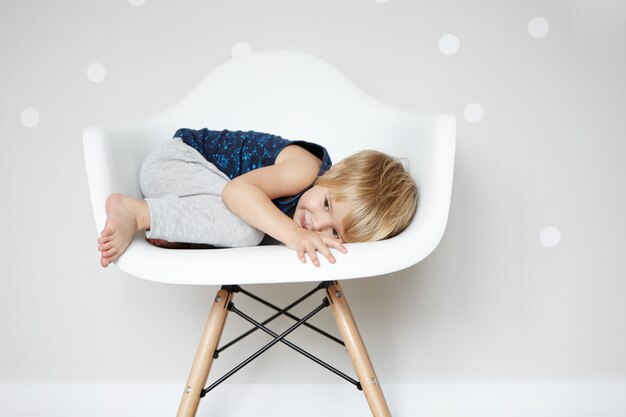Happy carefree childhood. Sweet Caucasian infant rolling himself up in white designer chair, hiding from his friends while playing hide-and-seek. Cute baby boy having fun indoors.