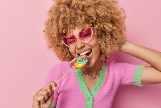 Happy carefree curly haired woman cannot live without sweets bites multicolred candy wears heart shaped sunglasses and t shirt isolated over pink background Taste rainbow Sweet life concept