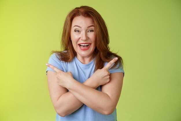 Happy carefree entushiastic redhead funny mature female having fun positive attitude cross arms body pointing sideways show left right products laughing happily like both choices green wall