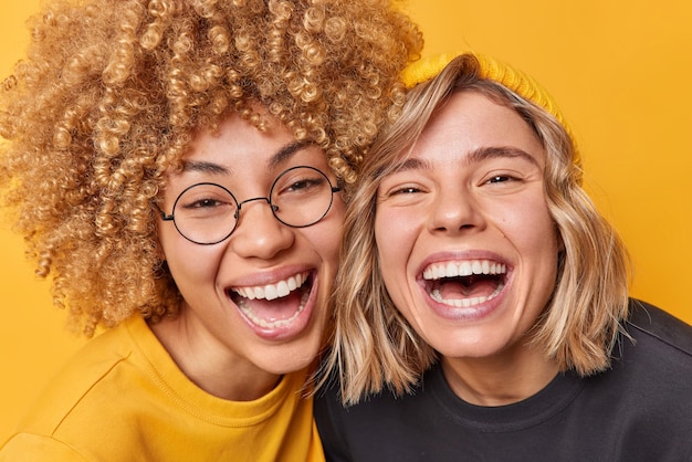 Happy carefree friendly female models laugh joyfully stand next to each other smile toothily have upbeat mood isolated over yellow background People friendship and positive emotions concept