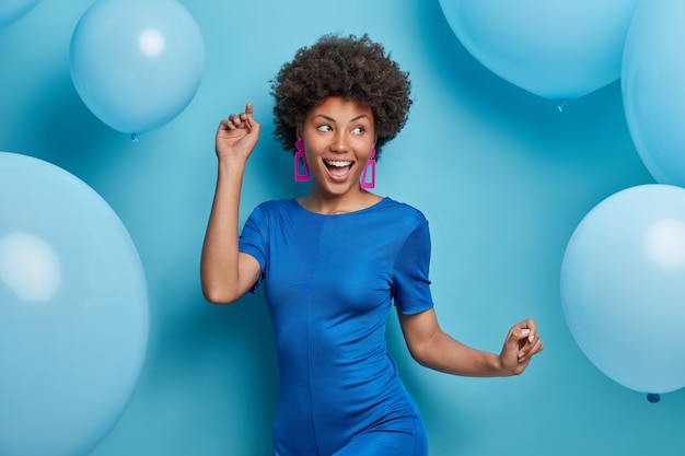 happy carefree woman dances and has fun dressed in fashionable clothes has festive mood poses against blue balloons
