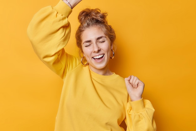Happy carefree woman makes triumph dance celebrates winning prize laughs gladfully keeps eyes closed dressed in casual sweatshirt moves against yellow background has fun hooray sweet success