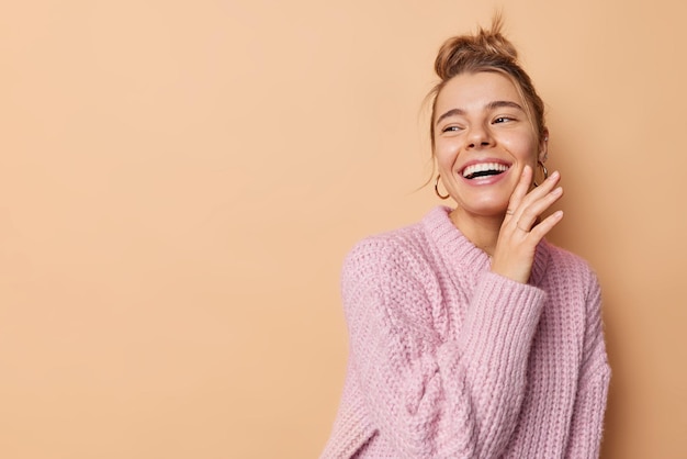Happy carefree woman with hair gathered in bun touches face gently looks away smiles broadly wears knitted sweater isolated over beige background blank copy space for your promotional content